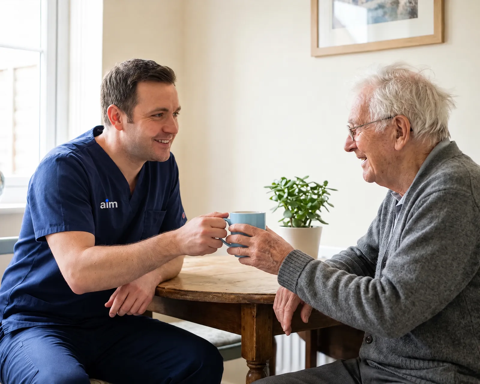 A carer in navy scrubs shares a cup of tea with a smiling older man at his kitchen table.